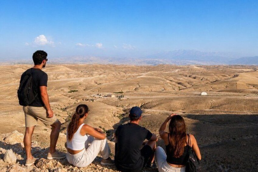 Un groupe de personnes est assis et se tient debout sur un terrain rocheux, d’où l’on peut admirer un vaste paysage aride et des montagnes lointaines. 3
