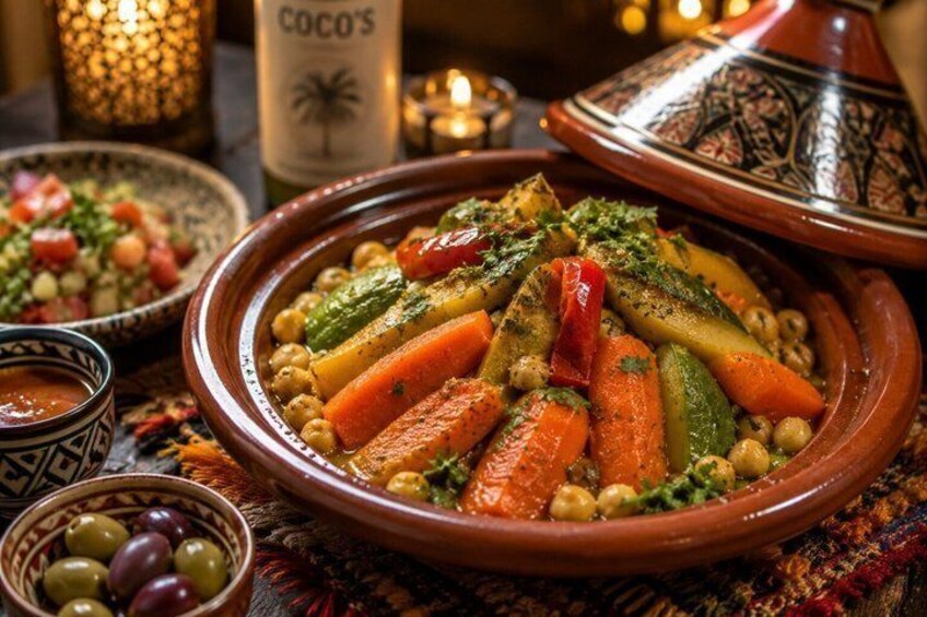 Un plat traditionnel marocain, un tajine, préparé avec des légumes et des pois chiches, accompagné d’un plat à tajine décoré, et une bouteille portant l’étiquette « COCO’S » en arrière-plan. 2