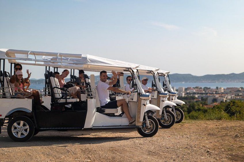 A white golf cart with passengers on a dirt road, overlooking a coastal cityscape. 2