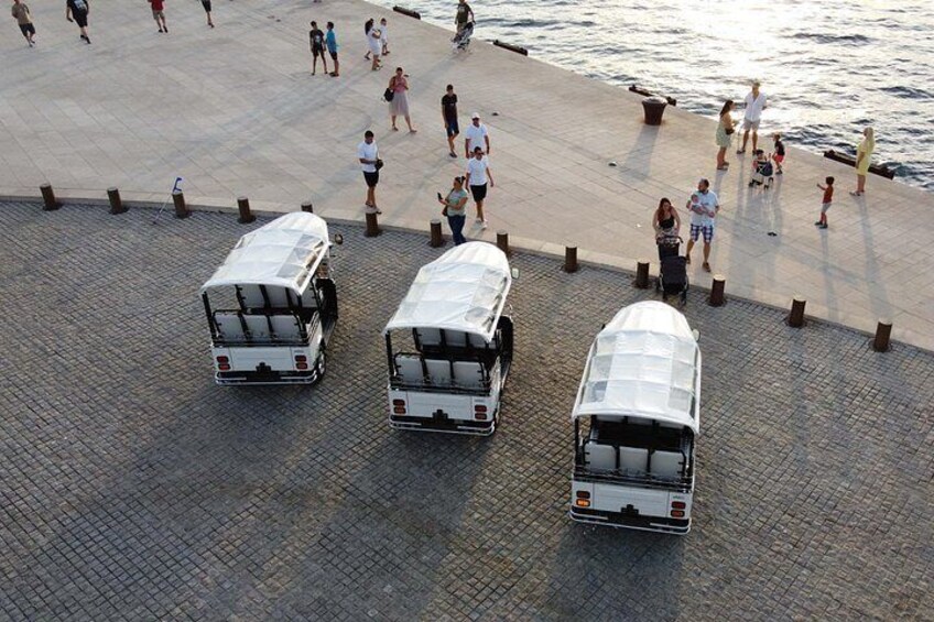 Three white buses parked on a paved area near a waterfront with people walking around. 2