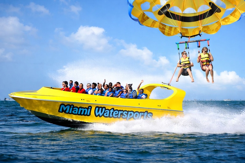 Keltainen Miami Watersports -vene, jossa on matkustajia, ja taustalla näkyy varjoliidossa leijaileva henkilö. 1