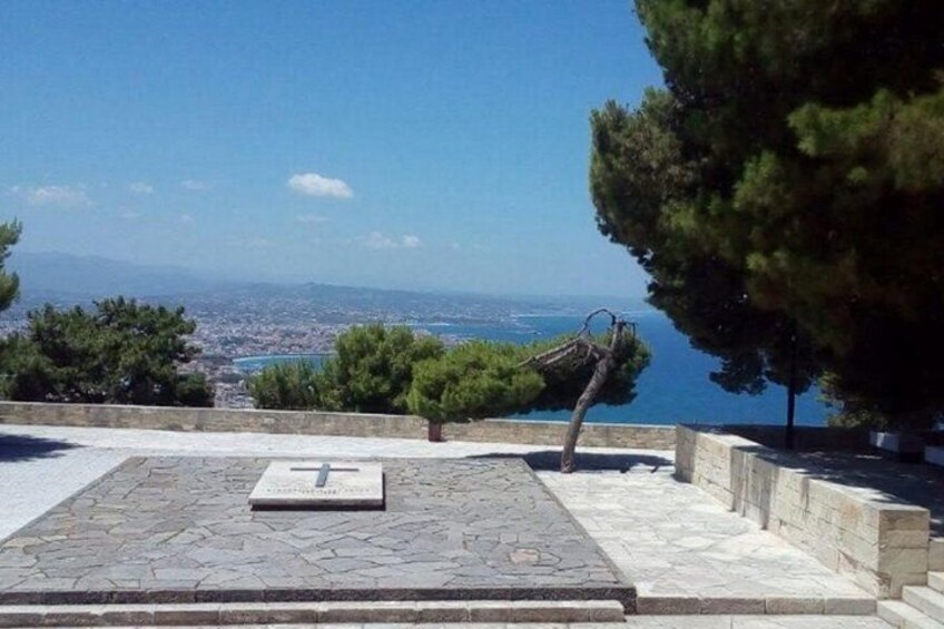 A stone-paved area with a bench and a sculpture, overlooking a coastal city and the sea. 2