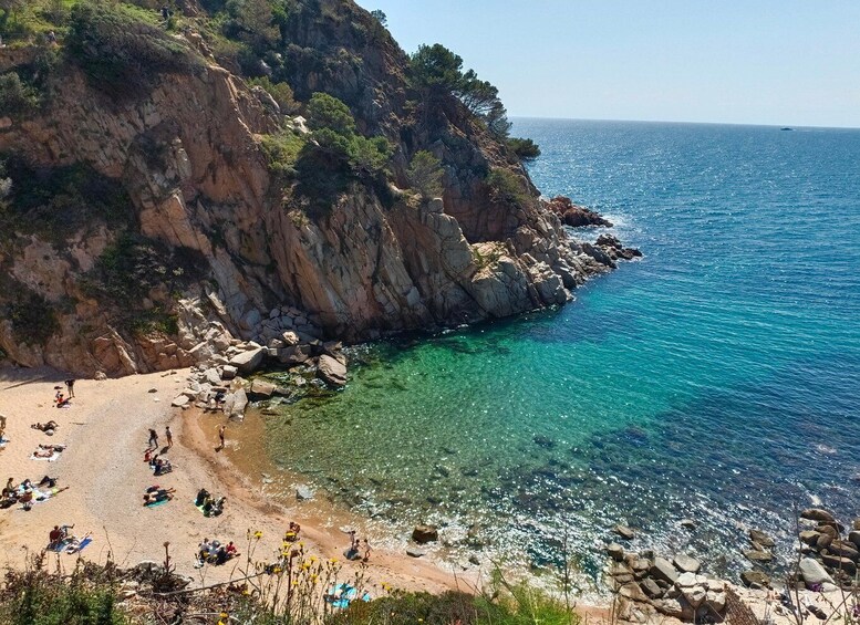 A rocky coastline with clear turquoise waters, a sandy beach, and people sunbathing. 2