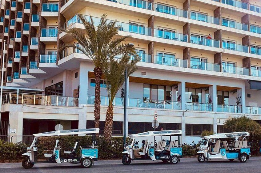 A multi-story hotel building with balconies, palm trees, and three tuk-tuks parked in front. 4