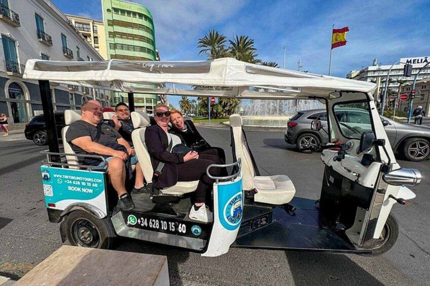 A golf cart with three passengers, parked on a street with buildings and palm trees in the background. 2