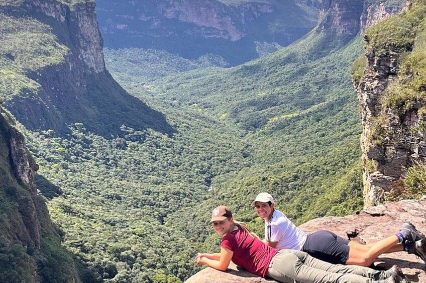 Two people sitting on a cliff edge overlooking a lush valley. 1