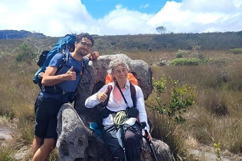 Two people sitting on a rock with backpacks, surrounded by grass and hills. 5