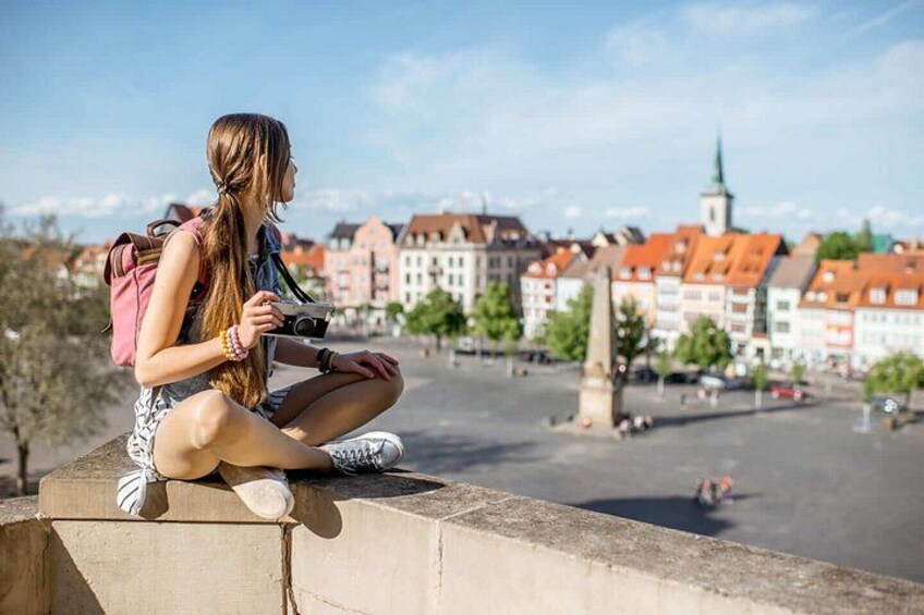 En person sidder på en udkragning og ser ned på en bytorv med bygninger og en kirke. 2