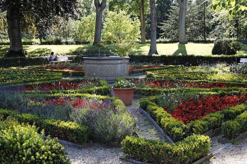 En velplejet have med forskellige blomsterbede, en central stenskulptur og en bænk i baggrunden. 4