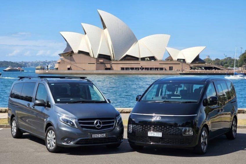 Two cars parked in front of the Sydney Opera House with the harbor in the background. 2