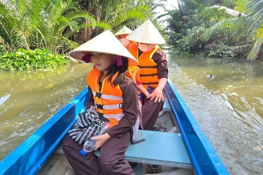 Deux personnes dans un bateau bleu, portant des gilets de sauvetage et des chapeaux coniques, entourées de verdure et d’eau. 1