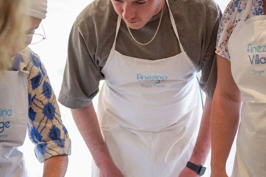 A person wearing a white apron with 'Anezia Dryos' printed on it, standing with their hands on their hips. 4