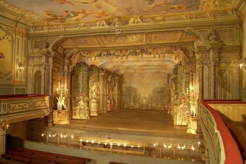 An ornate theater interior with a grand stage, elaborate ceiling frescoes, and classical columns. 5
