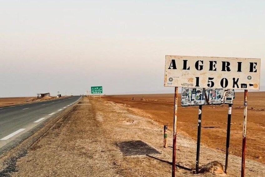 A road with a sign indicating 'ALGERIE 150Km' and a desert landscape. 5