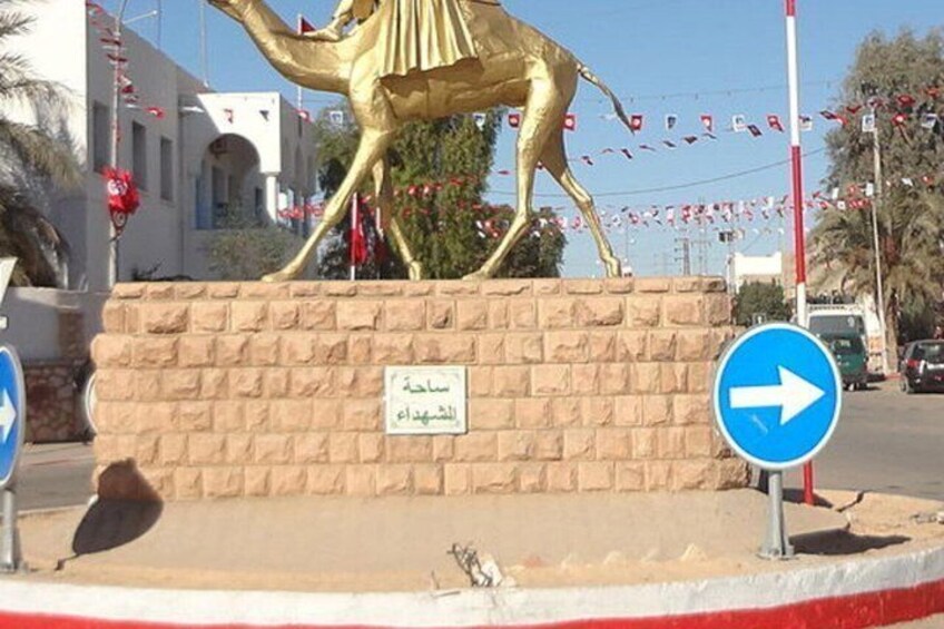 A golden camel statue on a brick pedestal with a blue directional sign and a bird on the ground. 2
