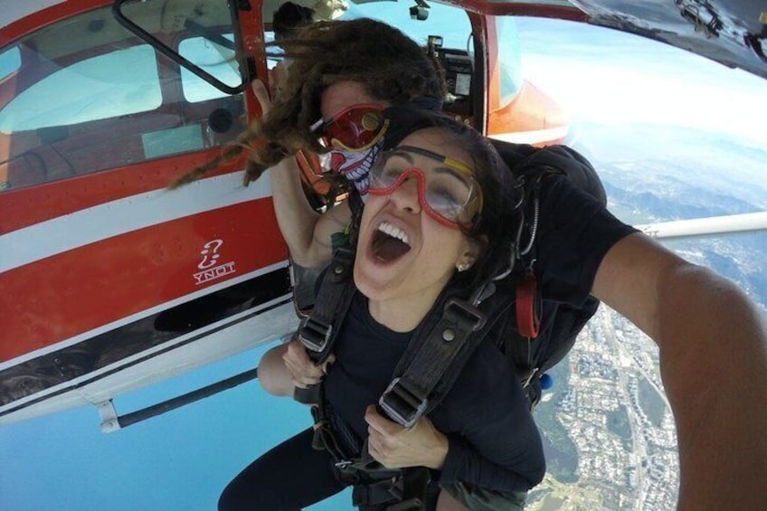 Eine Person im Harness, eine weitere Person in Fallschirmsprungposition mit Fallschirm, vor einem roten und weißen Flugzeug. 4