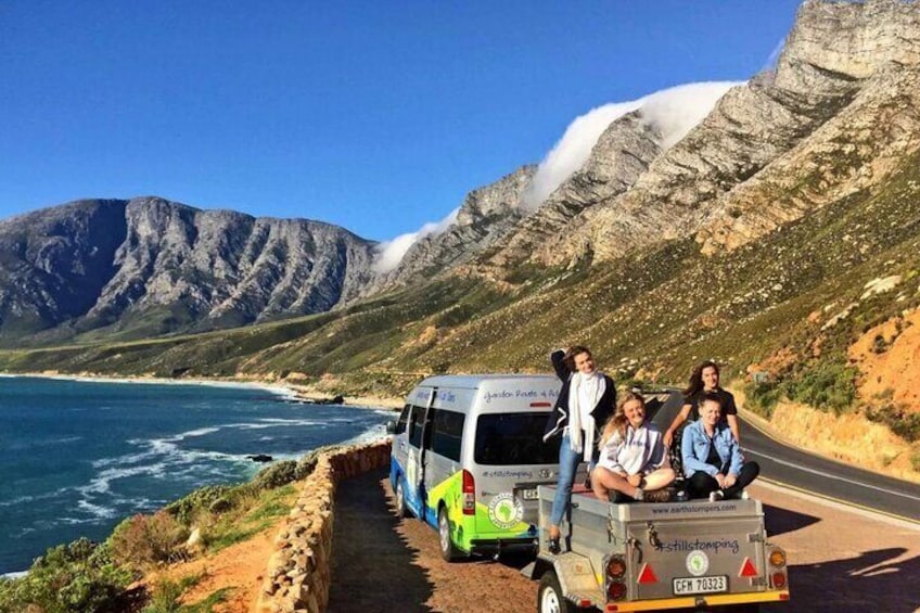 A group of people on a tour bus parked on a coastal road with mountains in the background. 1