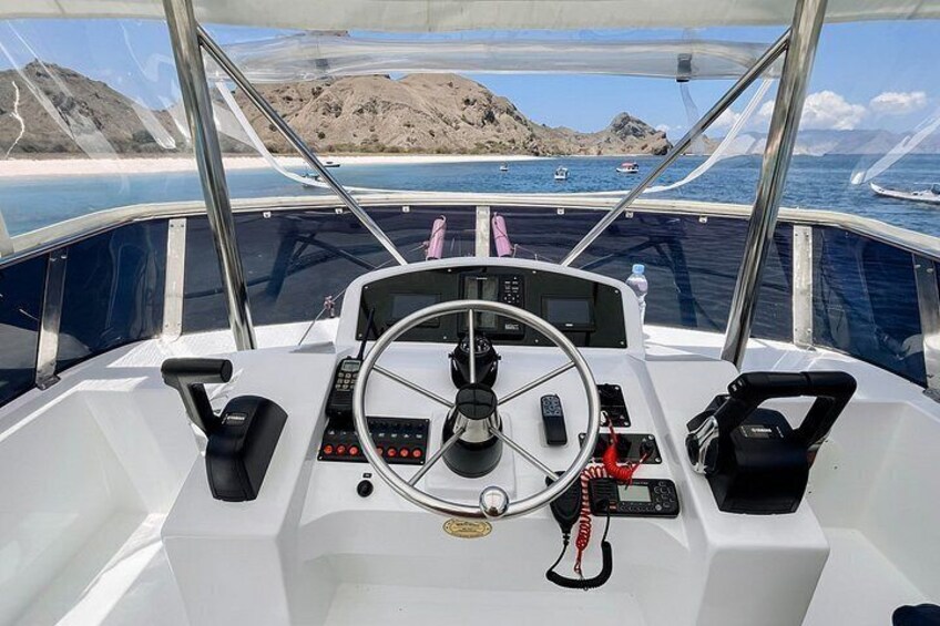 A boat's cockpit with a steering wheel, control panel, and a view of the sea and rocky landscape. 1