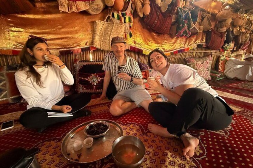 Three people sitting on a patterned carpet with a traditional setting, including a woven wall hanging and a metal tray with bowls. 5