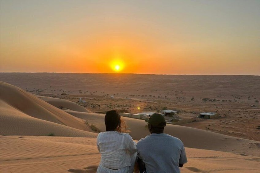 Two people overlooking a desert landscape with sand dunes and a sunset. 1
