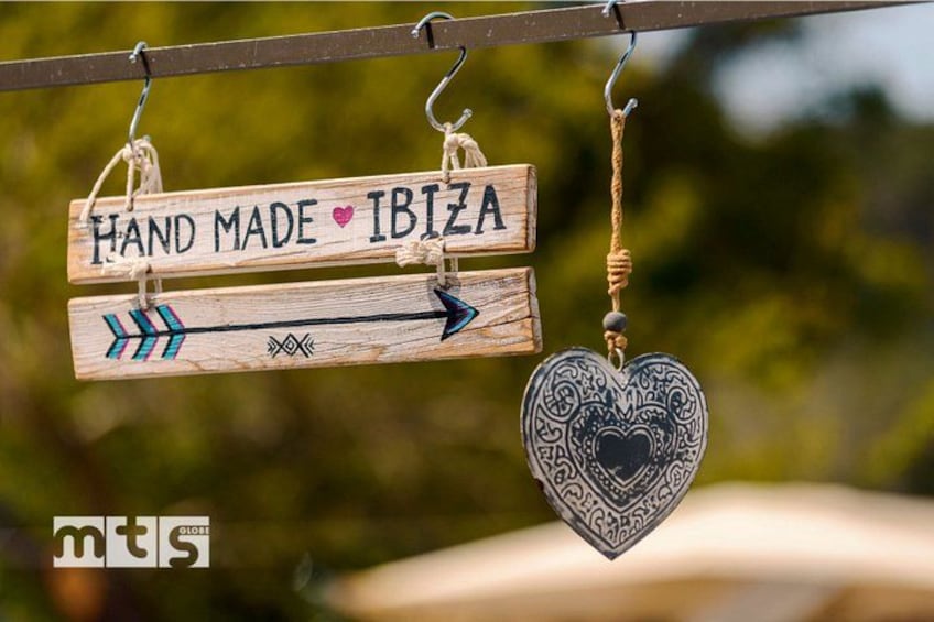 Een houten bord met 'HAND MADE IBIZA' en een naar rechts wijzende pijl, bevestigd aan een metalen stang. 2