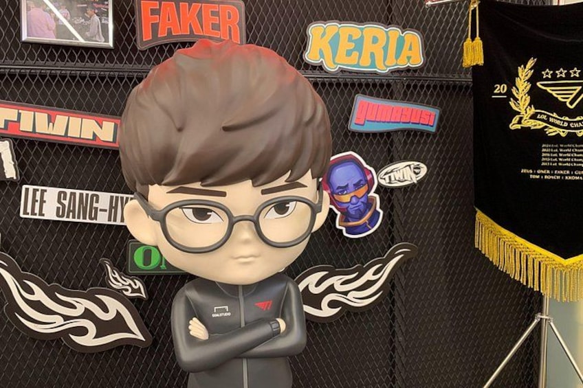 A figurine with a black jacket and glasses, standing in front of a backdrop with various logos and text, including 'FAKER' and 'KERIA'. 2