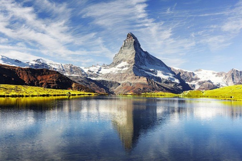 A mountain peak reflecting in a calm lake surrounded by green hills and snow-capped mountains. 5
