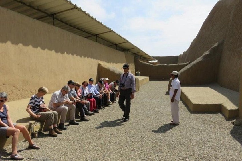 A group of people seated on benches in an outdoor setting with a man walking and another standing. 3
