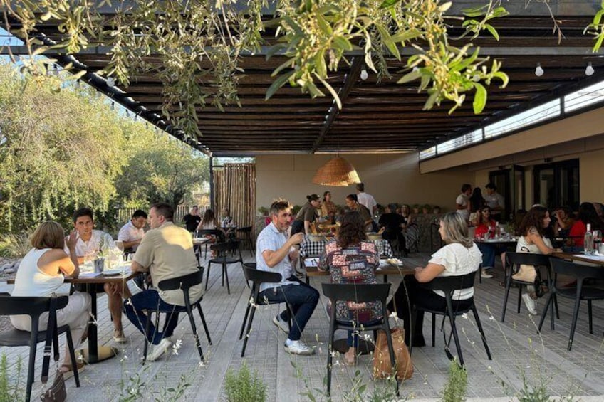 Ett utomhuskafé med människor som sitter och äter vid bord under en pergola. 2