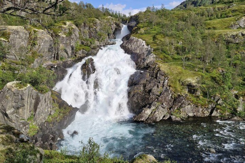 A waterfall cascading over rocks into a river, surrounded by greenery and hills. 3