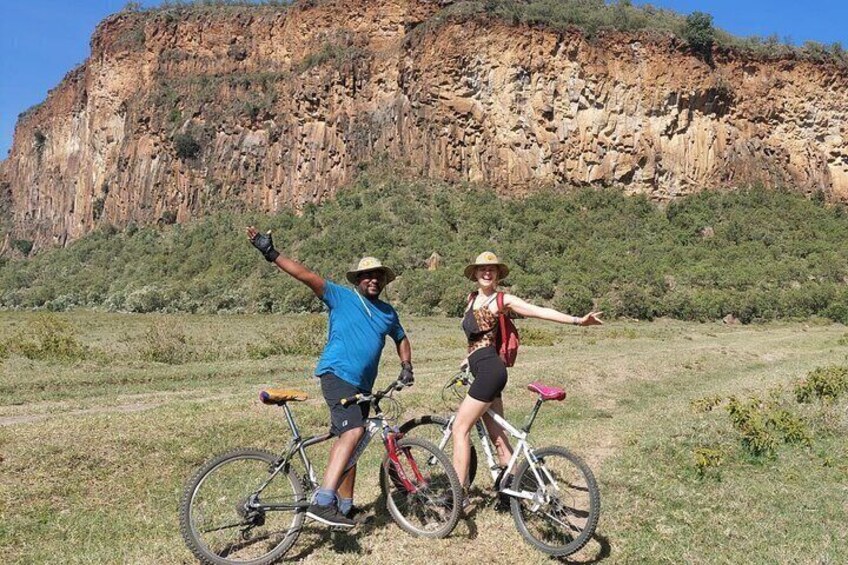 Two people on bicycles in a grassy field with a rocky cliff in the background. 5