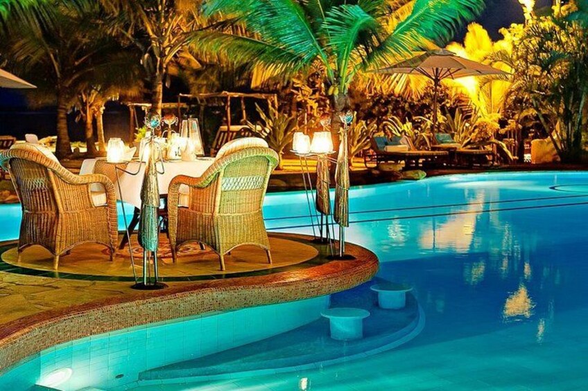 A poolside dining area with wicker chairs and a table set for two, surrounded by palm trees and a pool. 2
