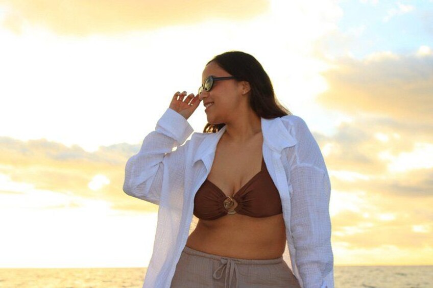 Una persona con camisa blanca y top de bikini marrón, de pie junto al mar con un atardecer de fondo. 1