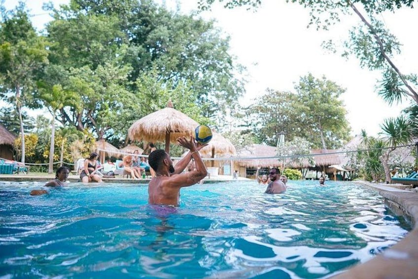 En swimmingpool där folk spelar volleyboll, omgiven av träd och stugor med halmtak. 3