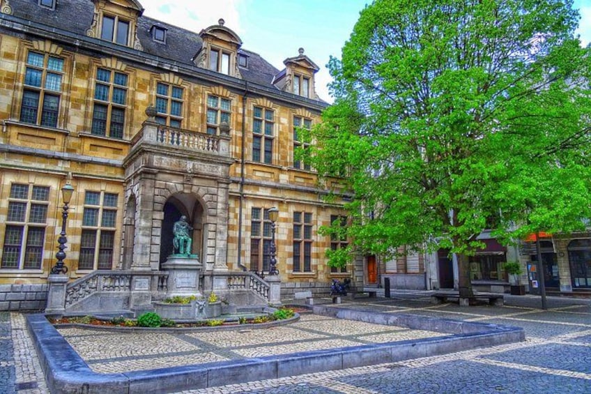 A historic building with a statue in front, a tree, and cobblestone pavement. 3