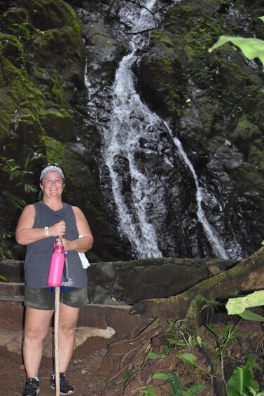 A person standing in front of a waterfall with a pink water bottle and a walking stick. 1
