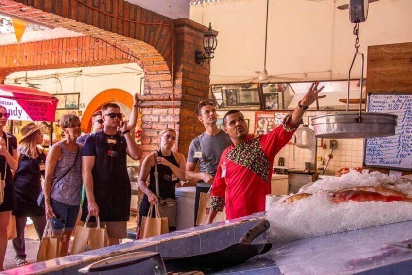 Un vendeur de nourriture de rue présente un poisson sur une table, des gens sont rassemblés autour, et un bâtiment en briques se trouve à l'arrière-plan. 1