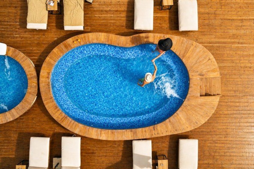Un terrazza in legno con tre piscine circolari, poltrone da relax bianche e un tavolino con un cappello. 3