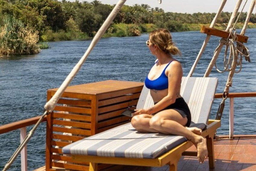 A person sitting on a wooden deck chair by a body of water, with a boat's railing and a rope visible. 2