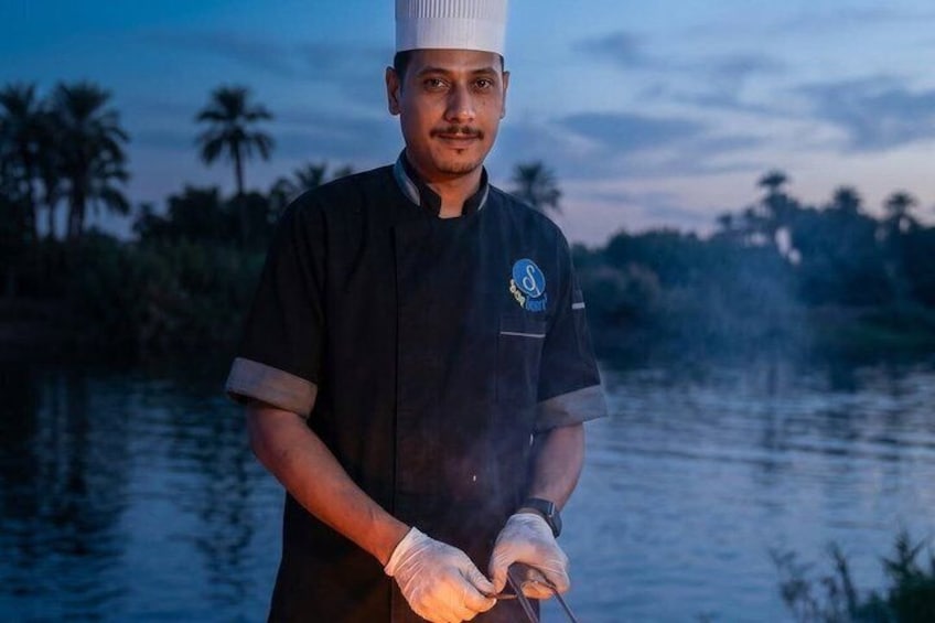 A chef in a black uniform with a white hat, standing by a body of water with palm trees in the background. 3