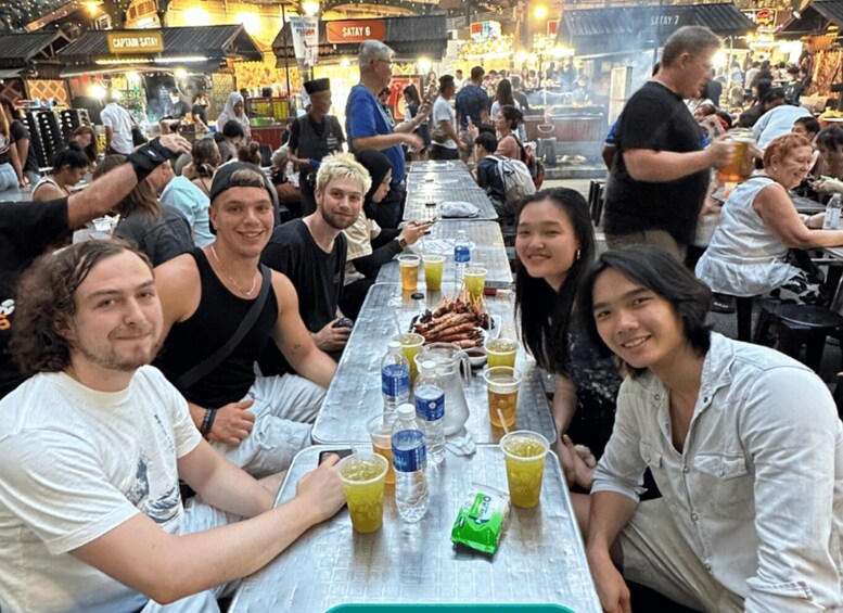 A group of people seated at a long table in a busy outdoor market with various food items and drinks. 1