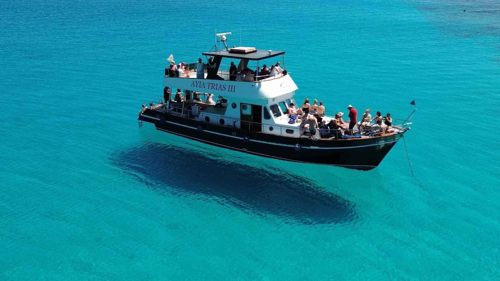 A boat named 'AYIA TRIAS III' with passengers on deck, floating on turquoise water. 5