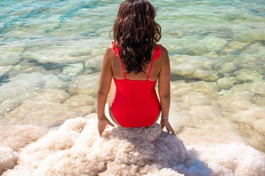 A person in a red swimsuit sitting on a white, porous rock formation by clear, shallow water. 2