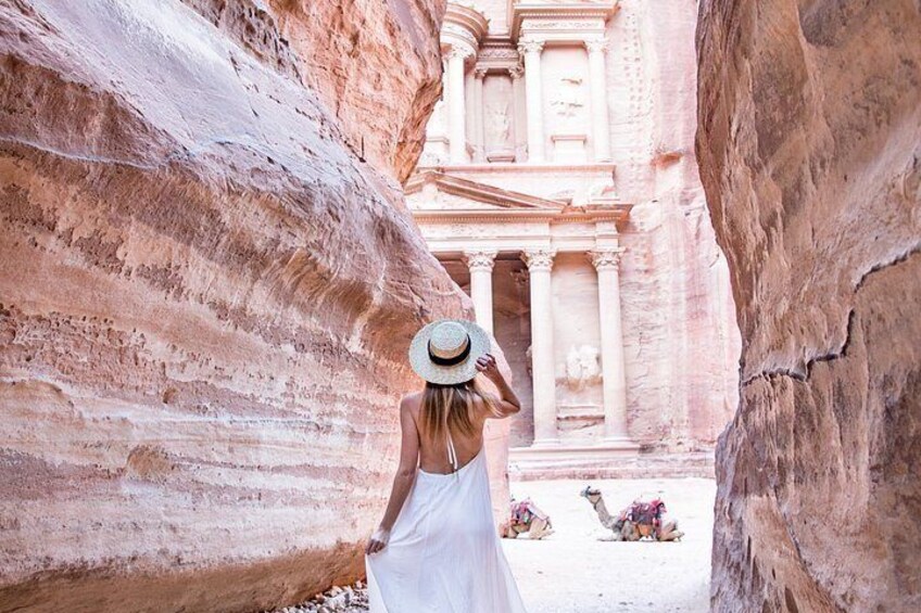 A person in a white dress and hat walking through a narrow canyon with ancient rock-carved structures in the background. 4