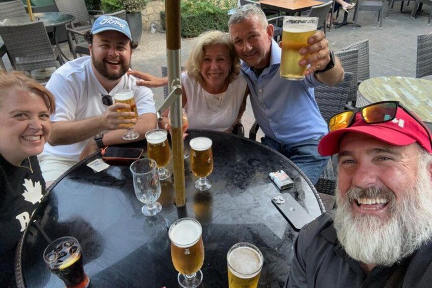 A group of people sitting around a round table with beer glasses, a grill, and outdoor furniture. 2