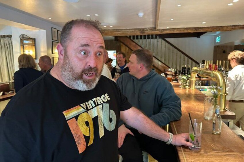 A man in a black shirt with a vintage design, a bar area with bottles and glasses, and a staircase in the background. 5