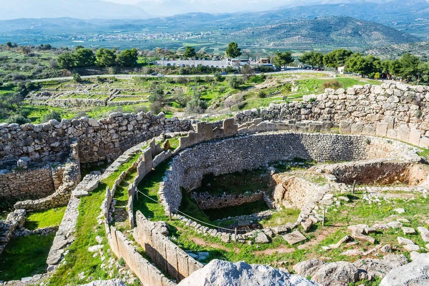 Ancient ruins with stone walls and a circular structure, set against a backdrop of green hills and distant mountains. 1