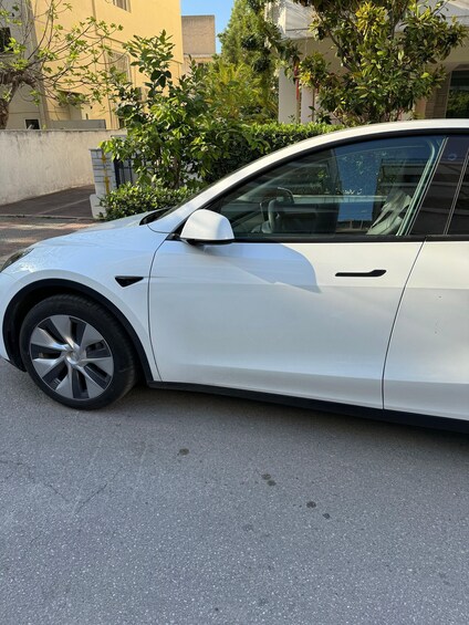 A white car parked on a paved road with buildings and greenery in the background. 2