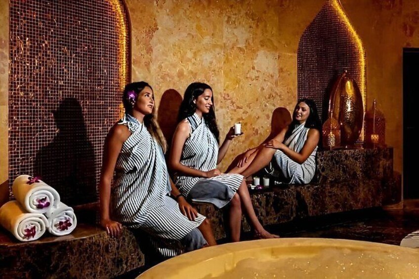 Three people in striped dresses sitting by a spa pool with a mosaic wall and a decorative fountain in the background. 1