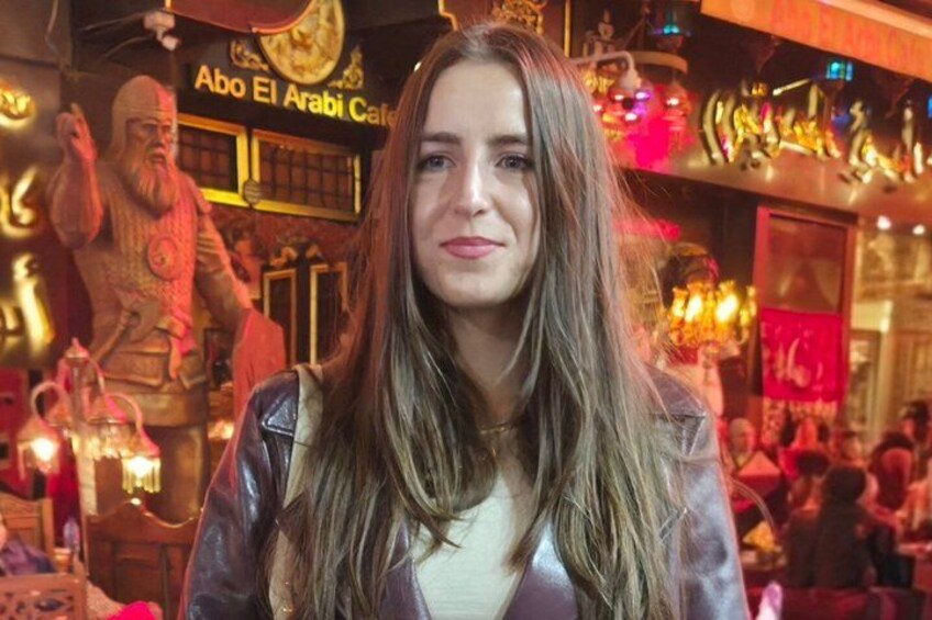 A person in a leather jacket at a restaurant with a sign reading 'Abo El Arabi Cafe' and a statue in the background. 5